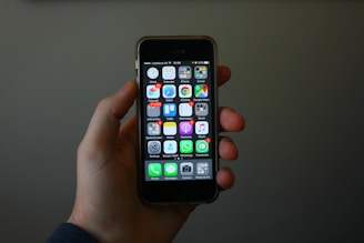 A hand is holding a smartphone displaying a home screen with various app icons. The apps include settings, phone, messages, and several others arranged in a grid pattern. The background is dimly lit, highlighting the phone and hand.