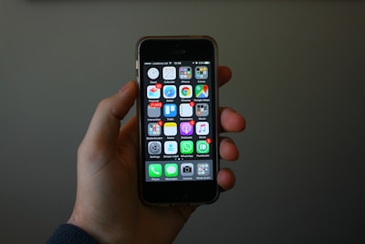 A hand is holding a smartphone displaying a home screen with various app icons. The apps include settings, phone, messages, and several others arranged in a grid pattern. The background is dimly lit, highlighting the phone and hand.