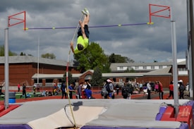 A person is in mid-air performing a pole vault over a high bar on an outdoor track field. They are wearing a bright yellow shirt and black shorts. Spectators and other athletes are visible in the background, with a school building and cloudy sky overhead.