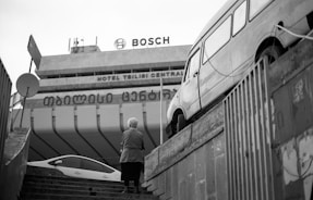 An urban scene featuring a staircase leading up to a large building with visible signage for Bosch and Hotel Tbilisi Central. A person with short hair stands on the steps facing away, while a vintage van is parked on a raised platform to the right. The scene is captured in black and white, adding a classic and timeless feel.
