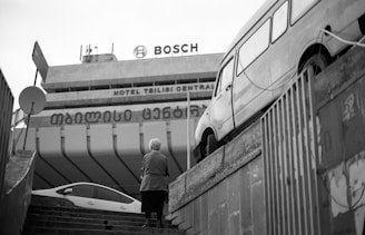 An urban scene featuring a staircase leading up to a large building with visible signage for Bosch and Hotel Tbilisi Central. A person with short hair stands on the steps facing away, while a vintage van is parked on a raised platform to the right. The scene is captured in black and white, adding a classic and timeless feel.