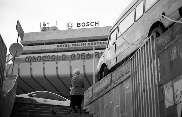 An urban scene featuring a staircase leading up to a large building with visible signage for Bosch and Hotel Tbilisi Central. A person with short hair stands on the steps facing away, while a vintage van is parked on a raised platform to the right. The scene is captured in black and white, adding a classic and timeless feel.