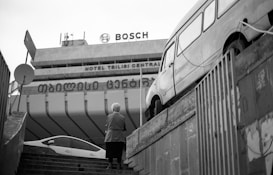 An urban scene featuring a staircase leading up to a large building with visible signage for Bosch and Hotel Tbilisi Central. A person with short hair stands on the steps facing away, while a vintage van is parked on a raised platform to the right. The scene is captured in black and white, adding a classic and timeless feel.