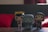 Three stylized figurines are positioned on a wooden surface with a dark blurred backdrop. The characters include two female figures and one with an oversized grin and green eyes resembling a cat. The background is dimly lit with red and dark tones.