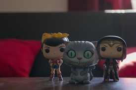 Three stylized figurines are positioned on a wooden surface with a dark blurred backdrop. The characters include two female figures and one with an oversized grin and green eyes resembling a cat. The background is dimly lit with red and dark tones.