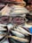 Various fresh fish and seafood are displayed on a market counter. Several types of fish, including silver and blue-toned fish, are arranged neatly in trays. Ice is used to keep the seafood fresh, and the setting appears to be a busy fish market with a variety of seafood offerings.