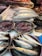 Various fresh fish and seafood are displayed on a market counter. Several types of fish, including silver and blue-toned fish, are arranged neatly in trays. Ice is used to keep the seafood fresh, and the setting appears to be a busy fish market with a variety of seafood offerings.