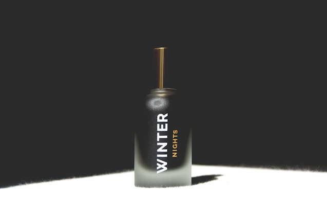 A perfume bottle with a sleek, dark-to-light gradient design and text reading 'Winter Nights'. The bottle features a golden cap and is set against a dark background with light highlighting its form.