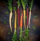 A variety of colorful carrots arranged in a line on a dark, textured background. The carrots have different colors, including white, yellow, orange, and purple, with green leafy tops still attached. Small carrot leaves are scattered around the arrangement.