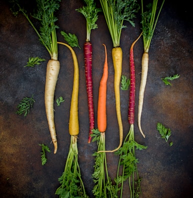 A variety of colorful carrots arranged in a line on a dark, textured background. The carrots have different colors, including white, yellow, orange, and purple, with green leafy tops still attached. Small carrot leaves are scattered around the arrangement.