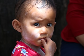 A young child with a worried expression, wearing a red floral-patterned outfit. The child is biting one finger and has some white powder on their face. The background is dark, with someone in a red shirt partially visible on the right side.