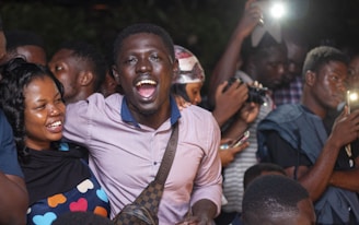 A group of people appear to be celebrating. A man in the center wearing a light pink shirt and carrying a crossbody bag is smiling widely. Beside him, a woman with long hair wearing a dark top with colorful heart patterns is also smiling. In the background, more people are visible, some holding cameras and phones, seemingly capturing the moment.