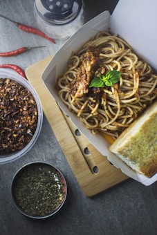 A takeout box filled with spaghetti topped with basil and a meat sauce is placed on a wooden cutting board. Beside it, there is a slice of garlic bread. Nearby, there are containers with dried chili flakes and herbs, as well as a few whole red chili peppers scattered on the table.