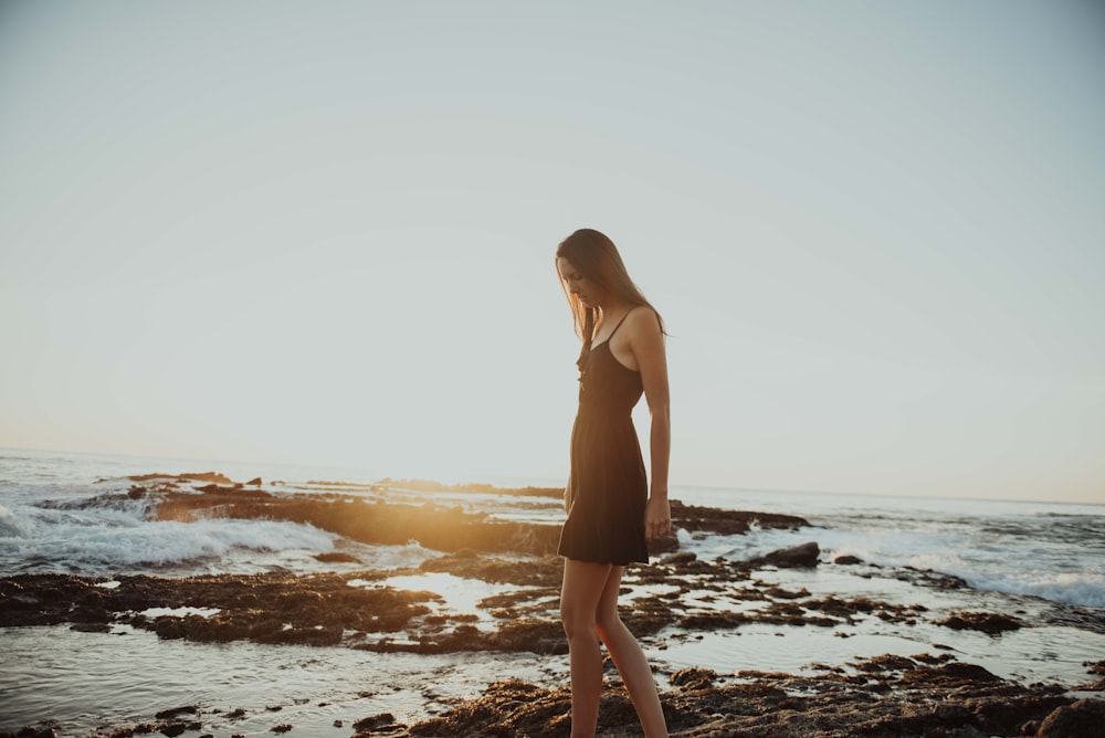 woman in black spaghetti strap dress walking on rock formation surrounded by body of water during daytime