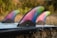A surfboard is equipped with three colorful fins, each displaying a gradient of vibrant colors including pink, blue, green, and purple. The surfboard sits on a grassy surface, with water droplets glistening on its surface. The background is slightly blurred, highlighting the bright and artistic design of the fins.