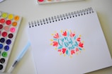 A spiral-bound drawing pad is open to a page displaying the word 'Contentment' in stylized, colorful typography surrounded by bright floral designs. Nearby, a watercolor paint set with various vibrant colors and a blue water brush pen are placed on a light-colored surface.