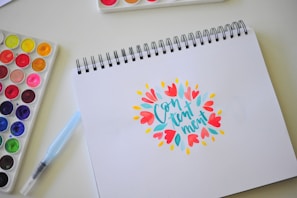 A spiral-bound drawing pad is open to a page displaying the word 'Contentment' in stylized, colorful typography surrounded by bright floral designs. Nearby, a watercolor paint set with various vibrant colors and a blue water brush pen are placed on a light-colored surface.