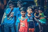 A group of six children are smiling and standing closely together in front of a vehicle. They are dressed in colorful casual clothing, and some are posing playfully. One child is holding a younger sibling on the right.