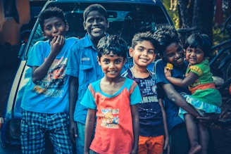 A group of six children are smiling and standing closely together in front of a vehicle. They are dressed in colorful casual clothing, and some are posing playfully. One child is holding a younger sibling on the right.