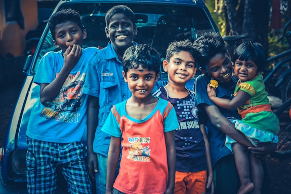 A group of six children are smiling and standing closely together in front of a vehicle. They are dressed in colorful casual clothing, and some are posing playfully. One child is holding a younger sibling on the right.