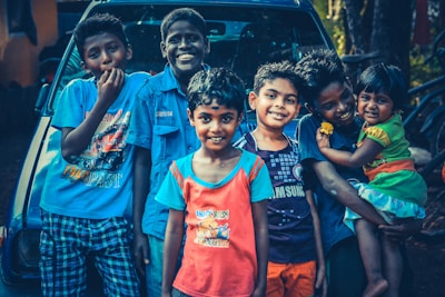 A group of six children are smiling and standing closely together in front of a vehicle. They are dressed in colorful casual clothing, and some are posing playfully. One child is holding a younger sibling on the right.
