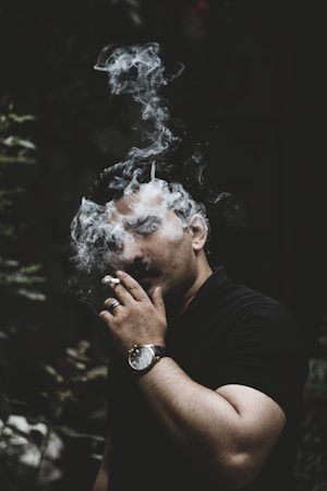 A person is standing with a cigarette in their hand, surrounded by swirling smoke. The scene is dimly lit, creating a moody and mysterious atmosphere. The individual is wearing a dark shirt and a watch, and their face is partially obscured by the smoke.