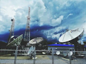 Large satellite dishes and telecommunications towers are set against a dramatic sky filled with dark clouds. A fenced area holds multiple satellite dishes of various sizes, with one prominently displaying the letters 'MRT'. Two tall communication towers rise in the background, indicating a broadcasting or data transmission facility.