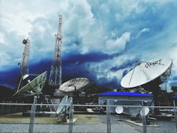 Large satellite dishes and telecommunications towers are set against a dramatic sky filled with dark clouds. A fenced area holds multiple satellite dishes of various sizes, with one prominently displaying the letters 'MRT'. Two tall communication towers rise in the background, indicating a broadcasting or data transmission facility.