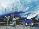 Large satellite dishes and telecommunications towers are set against a dramatic sky filled with dark clouds. A fenced area holds multiple satellite dishes of various sizes, with one prominently displaying the letters 'MRT'. Two tall communication towers rise in the background, indicating a broadcasting or data transmission facility.