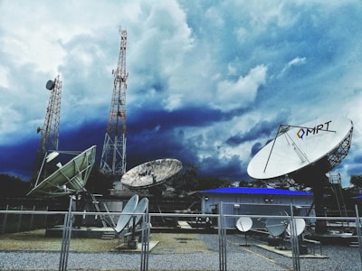 Large satellite dishes and telecommunications towers are set against a dramatic sky filled with dark clouds. A fenced area holds multiple satellite dishes of various sizes, with one prominently displaying the letters 'MRT'. Two tall communication towers rise in the background, indicating a broadcasting or data transmission facility.