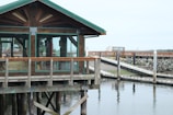 A wooden structure with large glass windows stands on stilts above the water, with a pier and rocky shoreline in the background. The structure features a green roof and wooden railings.