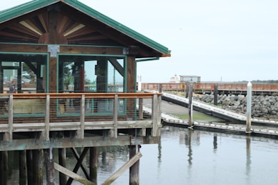 A wooden structure with large glass windows stands on stilts above the water, with a pier and rocky shoreline in the background. The structure features a green roof and wooden railings.