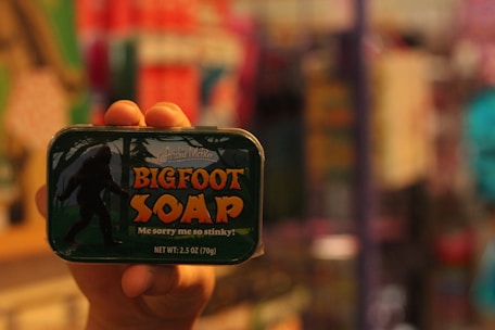 A hand holds a rectangular tin labeled 'Bigfoot Soap' with a silhouette of Bigfoot walking in a forest setting on it. The background is blurred, showing colorful shelves, possibly in a store.