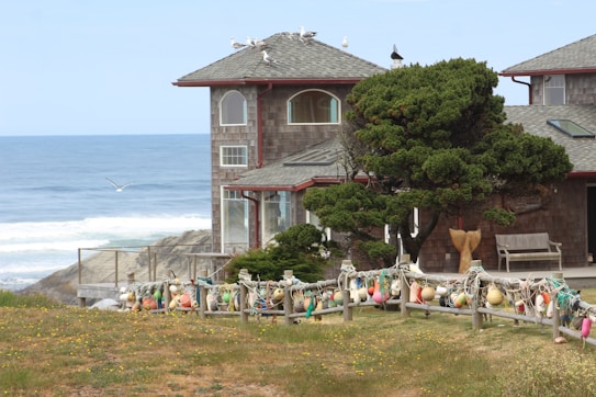 A coastal house with a gray shingled roof is surrounded by greenery and decorated with colorful buoys along a wooden fence. The ocean waves crash against the rocky shore in the background, while seagulls fly overhead and perch on the roof. A bench sits near a sculpted tree, adding to the serene atmosphere.