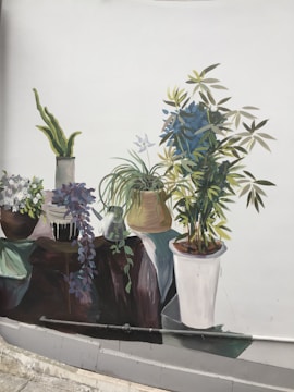 A mural depicts a collection of potted plants arranged on a surface. The plants vary in size and type, including leafy greens and flowering varieties. The colors are vibrant, with a mix of green, blue, and other natural hues. The background is plain, highlighting the detailed illustration of the plants.