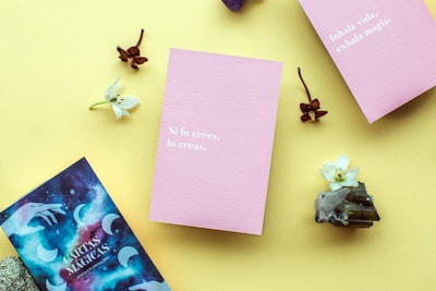 A layout on a yellow background featuring two pink cards with motivational phrases in Spanish, surrounded by small white flowers, brown dried flowers, crystals, and a cosmic-themed card.