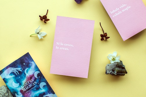 A layout on a yellow background featuring two pink cards with motivational phrases in Spanish, surrounded by small white flowers, brown dried flowers, crystals, and a cosmic-themed card.