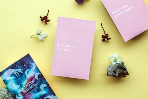 A layout on a yellow background featuring two pink cards with motivational phrases in Spanish, surrounded by small white flowers, brown dried flowers, crystals, and a cosmic-themed card.
