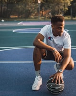A young man wearing a white t-shirt, black shorts, and patterned sneakers is crouching down on an outdoor basketball court. His right hand is resting on a black Nike basketball, and he appears focused, looking to his side. The background includes a basketball hoop and a court outlined in blue and purple.