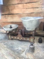 An antique balance scale rests on a stone surface. One side holds a small round weight and the other a large, empty basin. The scale has a rustic appearance with visible rust and an inscription on its base. In the background, wooden planks provide a rustic backdrop.
