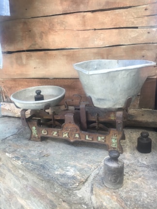 An antique balance scale rests on a stone surface. One side holds a small round weight and the other a large, empty basin. The scale has a rustic appearance with visible rust and an inscription on its base. In the background, wooden planks provide a rustic backdrop.