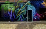 A vibrant street mural on a brick wall depicts a seated person wearing a hat and holding a flask, with swirling colors emanating from the flask. The artwork is rich in detail, showing bold movements in blue, green, purple, and yellow hues, creating a dynamic and fantastical scene.