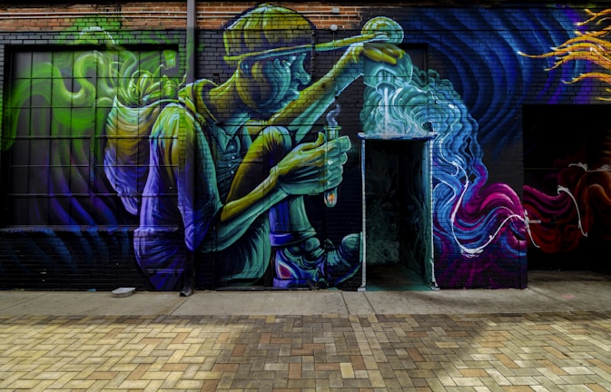 A vibrant street mural on a brick wall depicts a seated person wearing a hat and holding a flask, with swirling colors emanating from the flask. The artwork is rich in detail, showing bold movements in blue, green, purple, and yellow hues, creating a dynamic and fantastical scene.