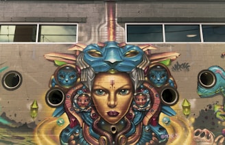A vibrant mural on a brick wall depicts a surreal, futuristic face with intricate details. The artwork features multiple faces, including ones resembling animals and mystical creatures, integrated into a complex design of colorful geometric shapes and patterns. The mural includes windows and circular elements that appear part of the building's architecture.