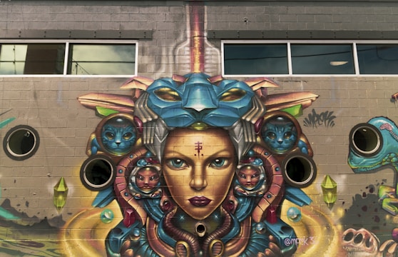 A vibrant mural on a brick wall depicts a surreal, futuristic face with intricate details. The artwork features multiple faces, including ones resembling animals and mystical creatures, integrated into a complex design of colorful geometric shapes and patterns. The mural includes windows and circular elements that appear part of the building's architecture.