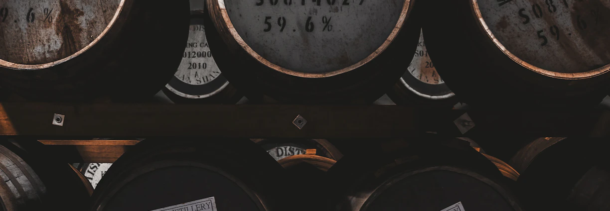 A close-up of aged whiskey barrels stacked in a dimly lit cellar, highlighting the wood grain and warm amber tones.