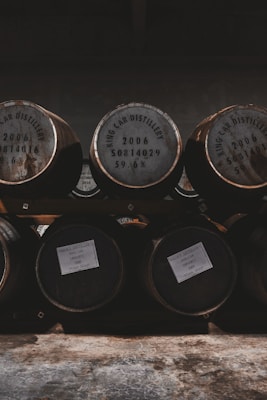 Several wooden barrels stacked in rows, with inscriptions from a distillery. The ambient light creates a warm, rustic atmosphere with shadows adding depth.