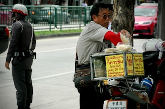 A street vendor is seen standing beside a motorcycle equipped with a food display box. The vendor appears to be selling snacks or packaged food, indicated by the items hanging on the motorcycle. A police officer in uniform is standing on the street, wearing a helmet and holding an object in one hand. The scene takes place on a city street, with a railing and moving vehicles in the background.