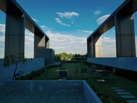 A rooftop garden with modern architectural elements featuring large rectangular frames extending into the sky. There are several tables and chairs scattered on the grassy area, surrounded by plants and minimalistic planters. The background reveals a partially cloudy blue sky.
