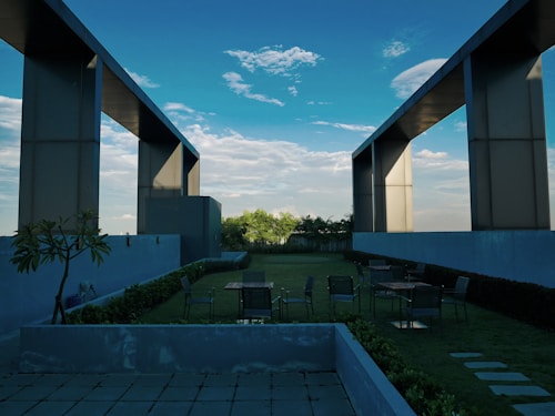A rooftop garden with modern architectural elements featuring large rectangular frames extending into the sky. There are several tables and chairs scattered on the grassy area, surrounded by plants and minimalistic planters. The background reveals a partially cloudy blue sky.